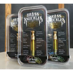 Three Brass Knuckles vape cartridge packages are displayed. The packages are labeled with different flavors: Girl Scout Cookie, Purple Haze, and Strawberry Cough. Each package contains a brass-colored vape cartridge.